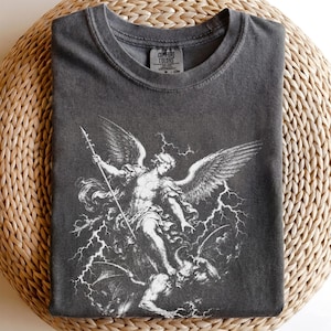 May include: A charcoal grey crew neck t-shirt featuring a white graphic print of an angel with wings holding a spear while standing over a horned figure amidst lightning bolts. The inner neck label reads Comfort Colors and includes size CM.