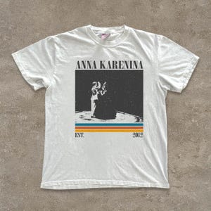 May include: White t-shirt featuring the text "ANNA KARENINA" above a graphic of two figures. Below the graphic are horizontal stripes in orange, blue, and yellow, with "EST. 2012" below the stripes. The shirt has a crew neck and short sleeves.