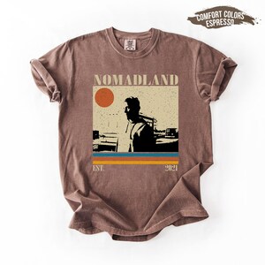 May include: A brown t-shirt with a graphic that says "NOMADLAND" above a silhouette of a person and a sun. Below the graphic are three horizontal stripes in blue, orange, and yellow. The text "EST. 2021" is below the stripes.
