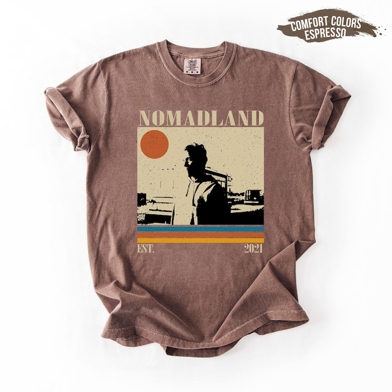 May include: A brown t-shirt with a graphic that says "NOMADLAND" above a silhouette of a person and a sun. Below the graphic are three horizontal stripes in blue, orange, and yellow. The text "EST. 2021" is below the stripes.