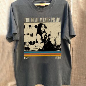 May include: Dark grey t-shirt with a graphic from the film "The Devil Wears Prada". The design features a black and white image of a woman, the film title, and the year 2006. The design has a retro style with horizontal stripes.