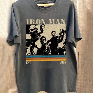 May include: Dark grey t-shirt with an Iron Man graphic. The design includes the text "IRON MAN" above a retro stripe design with the text "EST. 2008". The t-shirt is displayed on a wooden hanger.