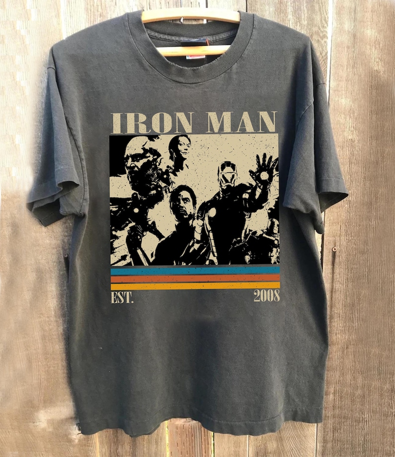 May include: Dark grey t-shirt with an Iron Man graphic. The design includes the text "IRON MAN" above a retro stripe design with the text "EST. 2008". The t-shirt is displayed on a wooden hanger.