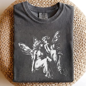 Può includere: Una t-shirt girocollo grigio antracite con una stampa grafica bianca di due figure alate sedute insieme. La maglietta presenta un'etichetta del marchio Comfort Colors all'interno del colletto. Il tessuto ha un aspetto slavato e testurizzato.
