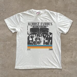 May include: White t-shirt featuring a graphic of a bus with the text "ALMOST FAMOUS" above a group of people. Below the bus is a horizontal stripe design in orange, blue, and yellow. The text "EST. 2000" is below the stripe.