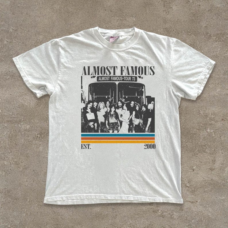 May include: White t-shirt featuring a graphic of a bus with the text "ALMOST FAMOUS" above a group of people. Below the bus is a horizontal stripe design in orange, blue, and yellow. The text "EST. 2000" is below the stripe.