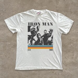 May include: White t-shirt with a black and white graphic of Iron Man characters. The design includes the text "IRON MAN" above the images, with "EST. 2008" below, and a horizontal stripe in orange, blue, and yellow.