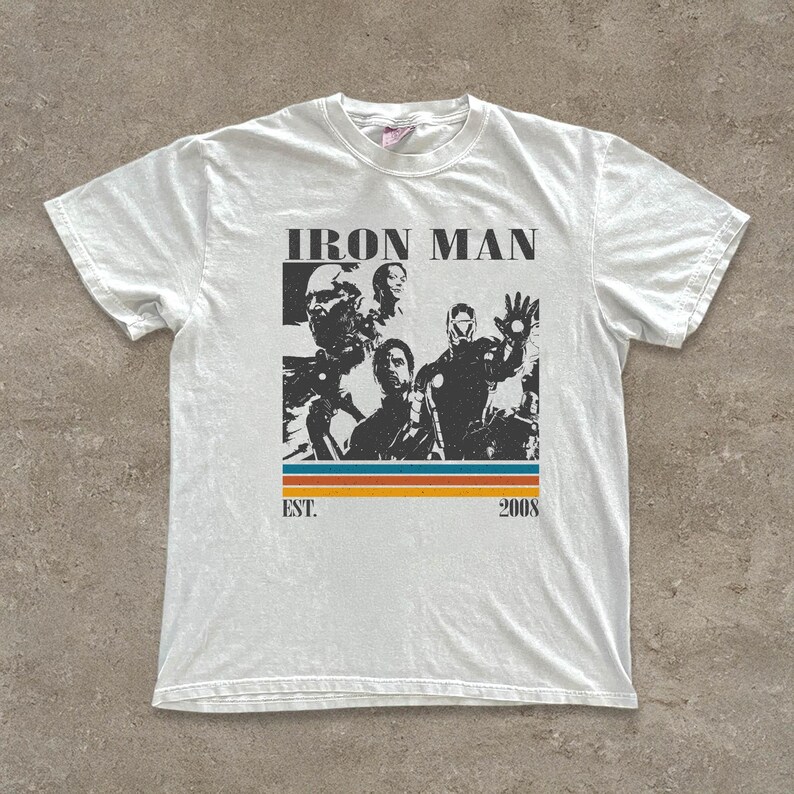 May include: White t-shirt with a black and white graphic of Iron Man characters. The design includes the text "IRON MAN" above the images, with "EST. 2008" below, and a horizontal stripe in orange, blue, and yellow.