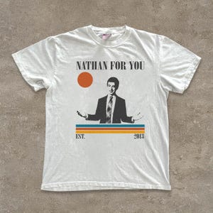 May include: White t-shirt featuring a graphic of a man in a suit with the text "NATHAN FOR YOU" above and "EST. 2013" below. The design includes an orange sun and horizontal stripes in blue, orange, and yellow.