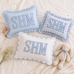 May include: Three rectangular throw pillows with ruffled edges. Pillow 1 is light blue with white polka dots. Pillow 2 is solid white with scalloped edges. Pillow 3 features a light blue and white gingham check. All display the letters SHM in floral.