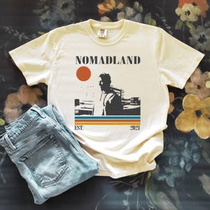 May include: Cream-colored t-shirt with the word "NOMADLAND" above a graphic of a person and a sunset. Below the graphic are the words "EST. 2021" in a retro style. Light blue denim jeans are folded next to the shirt.