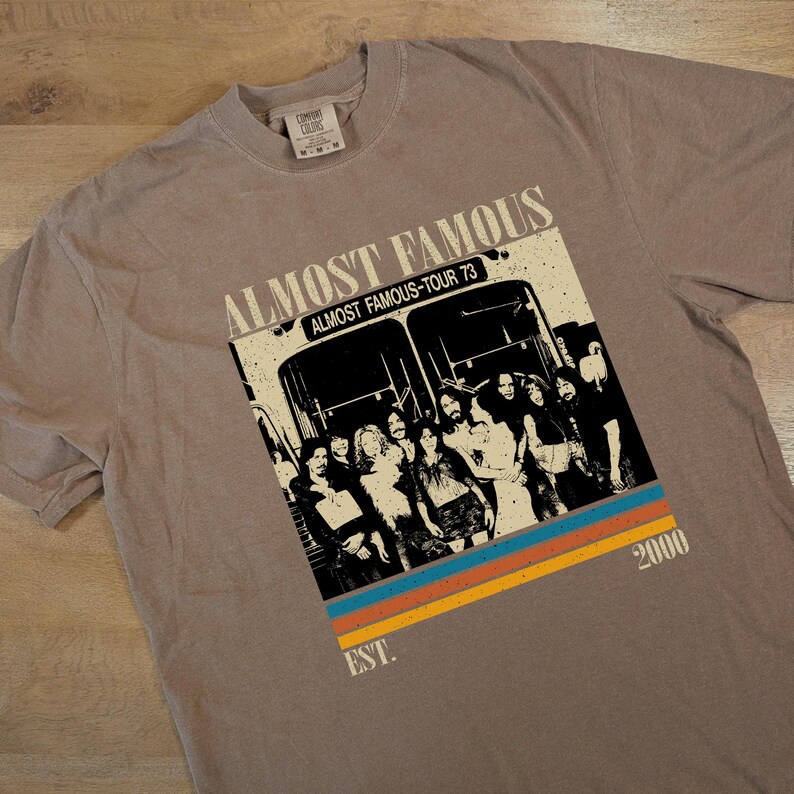May include: A taupe-colored t-shirt featuring a graphic from the movie "Almost Famous." The design includes the text "Almost Famous," "Almost Famous-Tour 73," and "2000." Below the bus image are three horizontal stripes in orange, blue, and yellow.