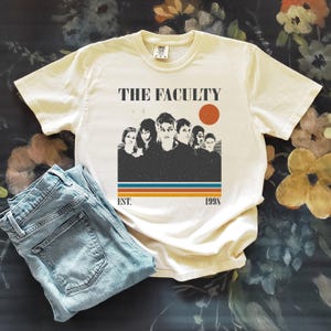 Może przedstawiać: Kremowa koszulka z napisem "THE FACULTY" nad grafiką przedstawiającą grupę osób i czerwone koło. Poniżej grafiki słowa "EST. 1998" w stylu retro. Jasnoniebieskie dżinsy są złożone obok koszulki.