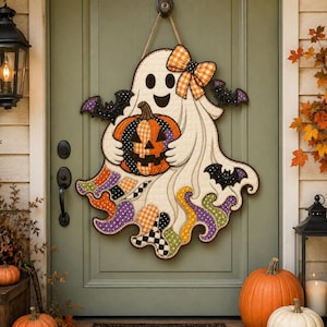May include: A quilted wall hanging shaped like a ghost holding a pumpkin, featuring a plaid bow, black bat accents, and a colorful patterned hem in orange, purple, green, and black, suspended by a rope from a green door.