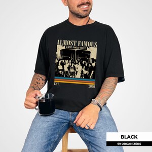 May include: Black t-shirt with a vintage-style graphic featuring the text "ALMOST FAMOUS" above a group of people in front of two buses. The graphic includes the text "ALMOST FAMOUS-TOUR 73" and "EST. 2000".