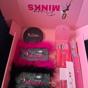 May include: A pink box labeled "Prissy Minks" contains beauty products. Items include a compact, eyelash applicators, lip gloss, and a pair of scissors in a pink case. The box is lined with pink and black feather trim.