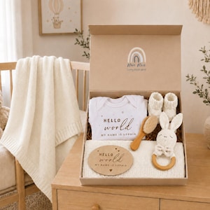 May include: A gift box containing a white baby bodysuit with the text Hello world MY NAME IS SOPHIA, a pair of white knit booties, a wooden hairbrush, a crochet rabbit rattle with a wooden ring, and a wooden plaque reading Hello world MY NAME IS SOPHIA.