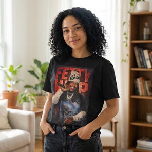 May include: Black t-shirt featuring a graphic of Fetty Wap holding a microphone, with the text "FETTY WAP" in red and white. The graphic includes a skull design. The person in the image is wearing the t-shirt and dark jeans.