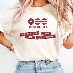 May include: A cream-colored short-sleeve t-shirt featuring a dark red glitter graphic. The design includes three circular icons, the text BTS WORLD TOUR, and two rectangular bars containing the names RM, JIN, SUGA, JIMIN, J-HOPE, and JUNGKOOK.