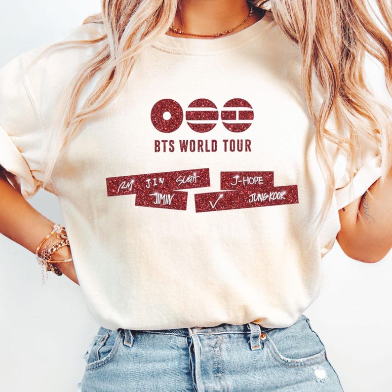 May include: A cream-colored short-sleeve t-shirt featuring a dark red glitter graphic. The design includes three circular icons, the text BTS WORLD TOUR, and two rectangular bars containing the names RM, JIN, SUGA, JIMIN, J-HOPE, and JUNGKOOK.