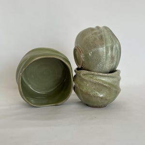 May include: Four handmade ceramic bowls in shades of green. One bowl is open and oval-shaped, while the other three are stacked and have a textured, rounded design. The bowls have a rustic, organic aesthetic, perfect for serving food or as decorative pieces.
