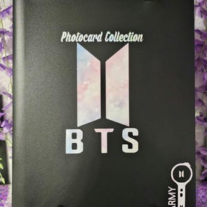 May include: Black photocard collection binder with the text "Photocard Collection" and "BTS" in white. The BTS logo is in the center with a pastel design. The binder also has an "ARMY" logo.