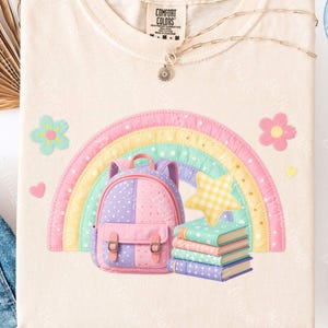 May include: A cream-colored t-shirt featuring a graphic print of a pastel-colored rainbow, a polka-dot backpack, a stack of four books, and a yellow gingham star. The shirt has a Comfort Colors brand label at the inner neckline.