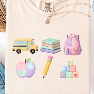 Puede incluir: Una camiseta de color crema con un estampado gráfico de temática escolar. El diseño incluye un autobús escolar amarillo, una pila de libros coloridos, una mochila rosa y morada, una manzana estampada, un lápiz amarillo y bloques de letras.