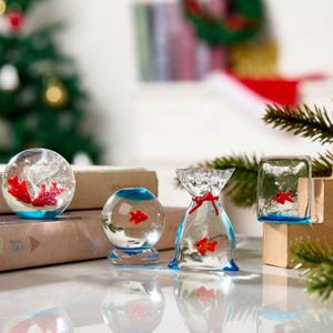 Peut inclure: Plusieurs ornements décoratifs en verre, chacun contenant un petit poisson rouge. Les ornements sont de formes variées : une sphère, un sac noué avec un ruban rouge et un cube. Les bases des ornements sont bleues. Un sapin de Noël est en arrière-plan.