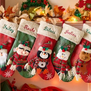 May include: Five Christmas stockings hang on a mantel. Each stocking is red and green with a white cuff and a name embroidered on it. The stockings are decorated with Santa, an elf, a penguin, a snowman, and a reindeer.