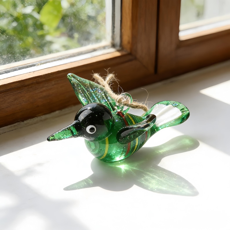Op de afbeelding: Een groene glazen vogelornament met een zwarte kop, wit oog en gestreept lichaam. De vogel heeft uitgespreide vleugels en is opgehangen aan een touwlus. Het ornament wordt in de buurt van een raam getoond.