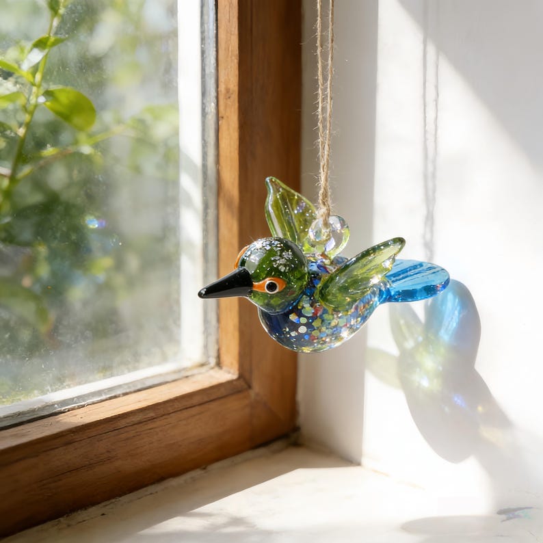 Op de afbeelding: Een kleurrijke glazen kolibrie-ornament met groene, blauwe en zilveren accenten. De vogel heeft een zwarte snavel en oranje ogen. Het hangt aan een touwtje, perfect voor een raam- of boomdecoratie.