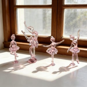 May include: Four pink glass ballerina figurines in different ballet poses. Each figurine wears a tutu. The figurines are displayed on a white surface in front of a wooden window frame.