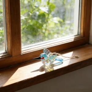 Op de afbeelding: Een helder glazen kolibrie-ornament met blauwe vleugels en lichaam, een zwarte snavel en rode accenten. De vogel is versierd met kleurrijke vlekken en heeft een touwtje om op te hangen. Het ligt op een houten vensterbank.