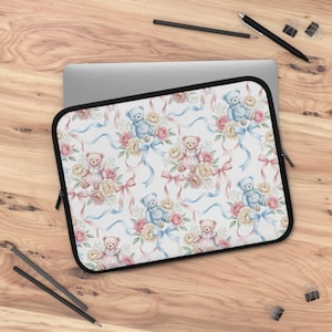 May include: Laptop sleeve featuring a repeating pattern of watercolor teddy bears, roses, and ribbons in pastel pink and blue hues. The sleeve has a black zipper and trim, and is set against a wooden surface.