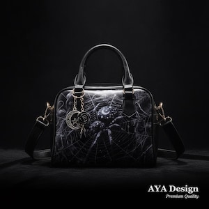 May include: A black handbag featuring a graphic print of a spider on a web with celestial moon and star motifs. The bag includes two top handles, a detachable shoulder strap, and a hanging charm with a crescent moon and a rose. Text reads AYA Design.