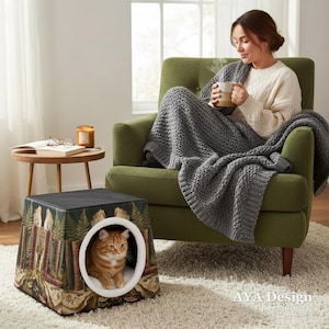 以下が含まれることがあります： 森をテーマにしたタペストリーデザインの正方形の猫用ベッド。 ベッドには白い内装の円形の開口部があり、オレンジ色のトラ猫が入っています。 ベッドは、緑のアームチェアの近くのラグの上にあります。