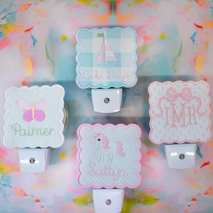 May include: Five decorative night lights with scalloped edges. Designs include a sailboat, butterfly, unicorn, and monogram. Each light has a unique embroidered design in pastel colors, with names or initials. The night lights are set against a colorful background.