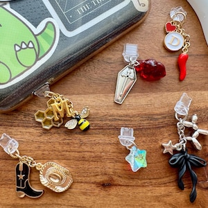 Peut inclure: Une collection de breloques décoratives pour téléphone avec divers motifs. Les breloques comprennent un nid d'abeille doré avec la lettre "M" et une abeille, une botte et un chapeau de cowboy noirs, un cercueil, un cœur rouge, un piment, une étoile, un chien ballon et un nœud noir.