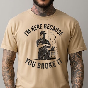 Può includere: T-shirt color cachi con la scritta nera "I'M HERE BECAUSE YOU BROKE IT". La grafica presenta una sagoma e un elettrodomestico rotto con fumo. La maglietta ha maniche corte e girocollo.