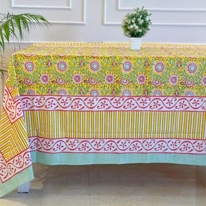 May include: A yellow floral tablecloth with a repeating pattern of pink flowers and green leaves. The tablecloth has a striped border in yellow and white, and a light green trim. A small potted plant sits on the table.