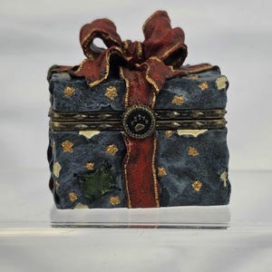 May include: A decorative box shaped like a gift, with a dark blue background and gold star accents. The box is adorned with a red ribbon and bow, and a paw print emblem. The box is a charming decorative item.