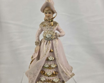 May include: A porcelain figurine of a woman in a pink dress and hat, adorned with gold accents. The figurine holds a bouquet and has intricate detailing on the dress and accessories. The figure is approximately 7 inches tall.