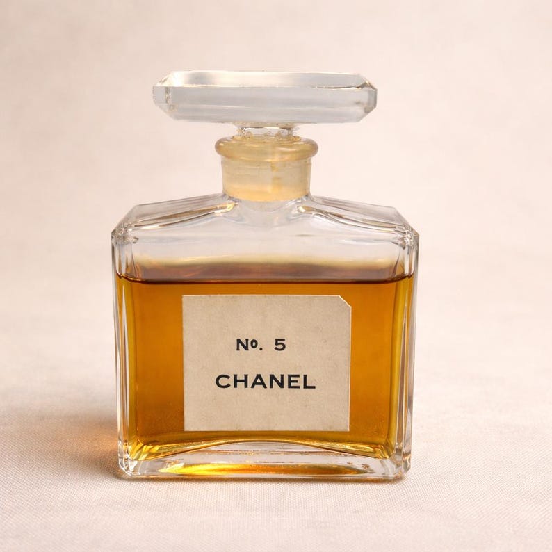 K&ouml;nnte beinhalten: Eine Vintage-Parf&uuml;mflasche mit klarem, geometrischem Design. Die Flasche enth&auml;lt eine goldbraune Fl&uuml;ssigkeit und tr&auml;gt ein Etikett mit der Aufschrift "No. 5 CHANEL". Der Verschluss ist rechteckig und klar. Die Gesamt&auml;sthetik ist klassisch und elegant.