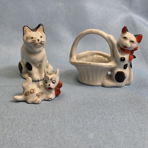 May include: Three ceramic cat figurines. One is sitting, white with black spots. Another is lying down, white with brown spots and a red bow. The third is a cat holding a basket, white with black spots and a red scarf.