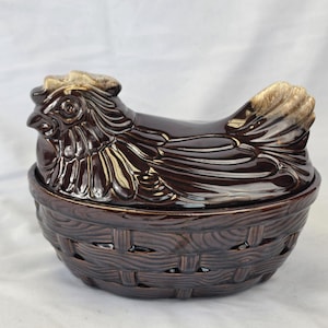 May include: A brown ceramic hen-shaped covered dish. The hen's head and body are detailed with feathers and a comb. The base is a basket weave design. The dish is a decorative kitchen item.