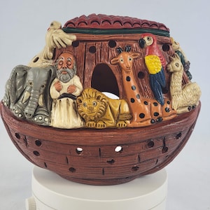 May include: A ceramic Noah's Ark figurine with a brown base and red roof. The ark features relief sculptures of animals, including an elephant, giraffe, lion, and parrot. The figurine is designed with cut-out holes for light.