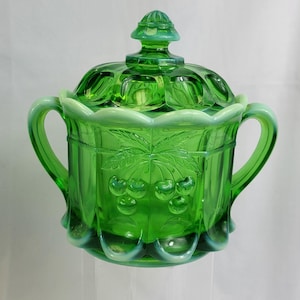 May include: Green glass sugar bowl with lid. The bowl features a detailed cherry design and two handles. The lid has a decorative finial. The glass has a slight opalescent effect around the edges.