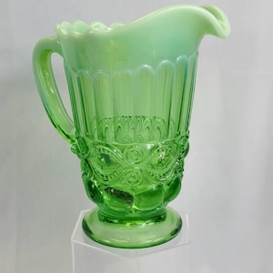 May include: A vintage green glass pitcher with a handle and a decorative base. The pitcher has a ruffled rim and vertical lines on the body. The base features an embossed floral design. The pitcher is displayed on a clear stand.
