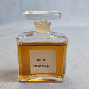K&ouml;nnte beinhalten: Eine Vintage-Parf&uuml;mflasche Chanel No. 5. Die klare Glasflasche hat eine rechteckige Form mit einem durchsichtigen Verschluss. Auf dem Etikett steht "No. 5 CHANEL". Die Fl&uuml;ssigkeit im Inneren ist bernsteinfarben.
